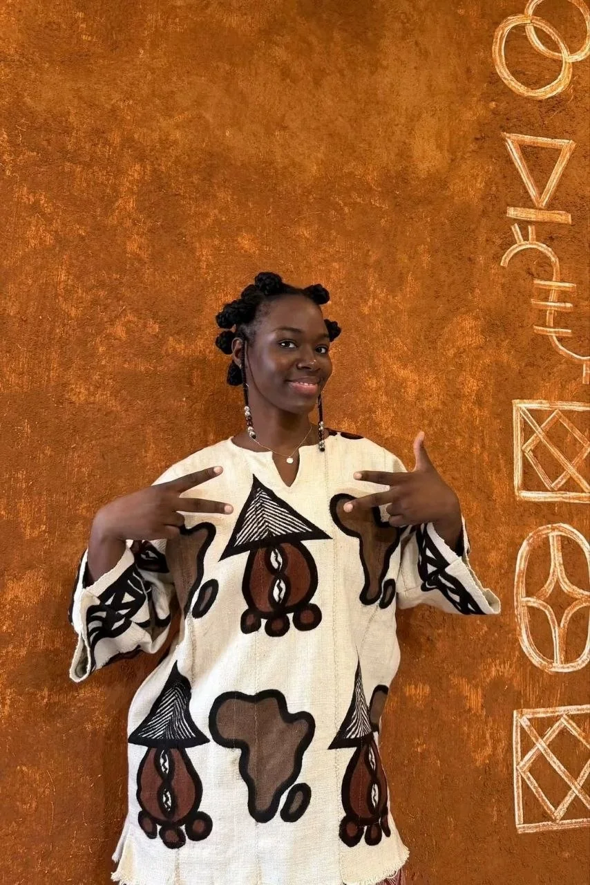 Une cliente de Kignon en chemise bogolan écrue, devant le mur ocre où sont peints les signes du logo